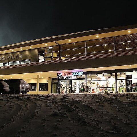 Ein modernes Sportgeschäft mit großen Fensterfronten bei Nacht. Der Eingang ist festlich beleuchtet und der Boden ist mit Schnee bedeckt.