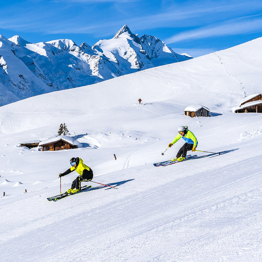 Das Bild zeigt eine winterliche Skiszene in den Bergen. Im Vordergrund fahren zwei Skifahrer parallel die Piste hinunter. Beide tragen gelbe Skijacken und dunkle Hosen. Sie sind in einer dynamischen Position, während sie die Abfahrt genießen, wobei ihre Skistöcke nach hinten zeigen. Die schneebedeckte Piste ist gut präpariert und glatt.  Im Hintergrund erhebt sich eine majestätische Berglandschaft mit schneebedeckten Gipfeln. Ein hoher, markanter Berg, möglicherweise ein Gletscher, ragt imposant in den strahlend blauen Himmel. Es gibt einige kleine, traditionelle Berghütten im oberen Bereich der Piste, die aus Holz gebaut sind und eine alpine Atmosphäre schaffen.  Die Szene vermittelt eine ruhige und klare Winterstimmung, geprägt von hellem Sonnenschein und unberührtem Schnee.